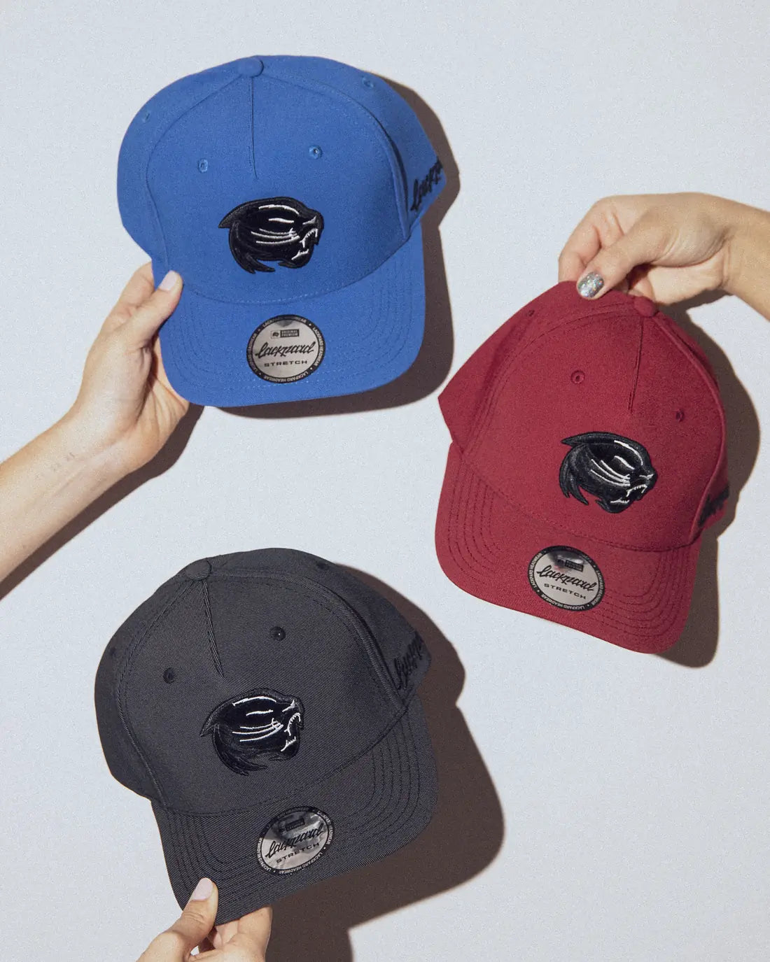 Three hands holding Lackpard caps in blue, burgundy and charcoal, each with a leopard patch.